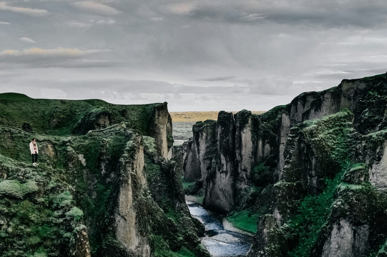 Person standing on green cliffs overlooking dramatic canyon landscape under cloudy sky